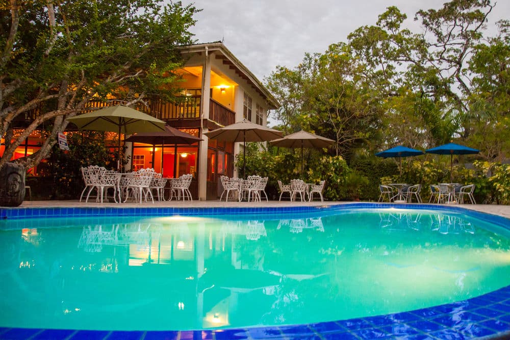 A tranquil pool scene with a house and outdoor seating surrounded by lush greenery.