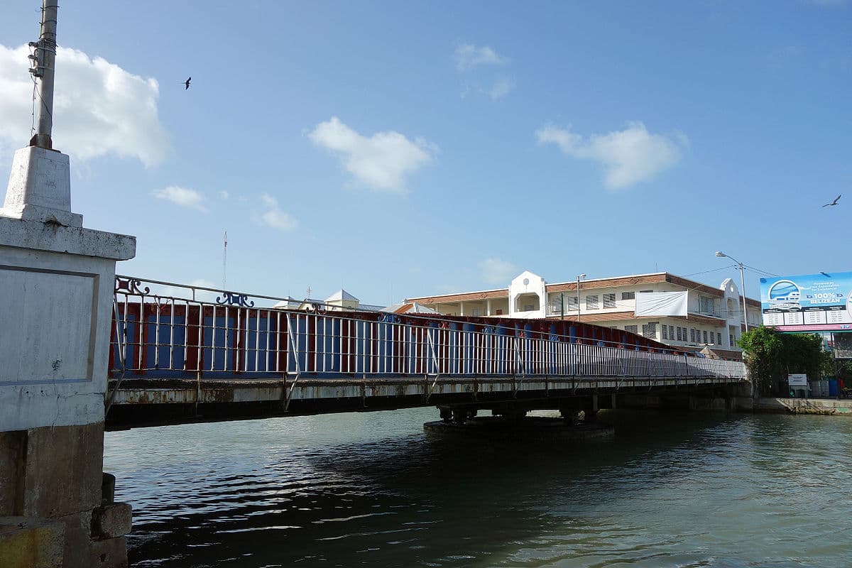 A bridge spans over calm waters, with buildings in the background under a clear blue sky.