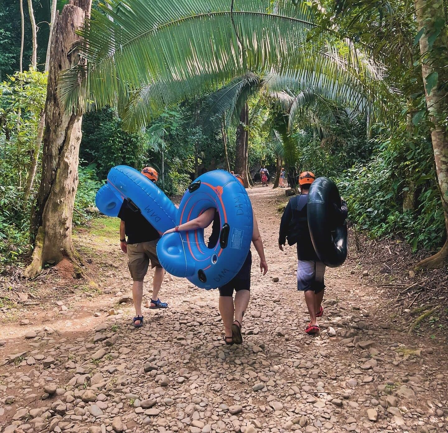 Three individuals walk down a rocky path in a lush forest, each carrying inflatable tubes.