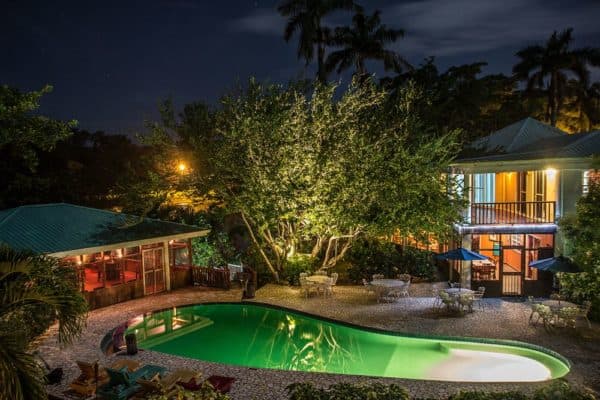 A well-lit pool area surrounded by lush trees and a cozy building at night.