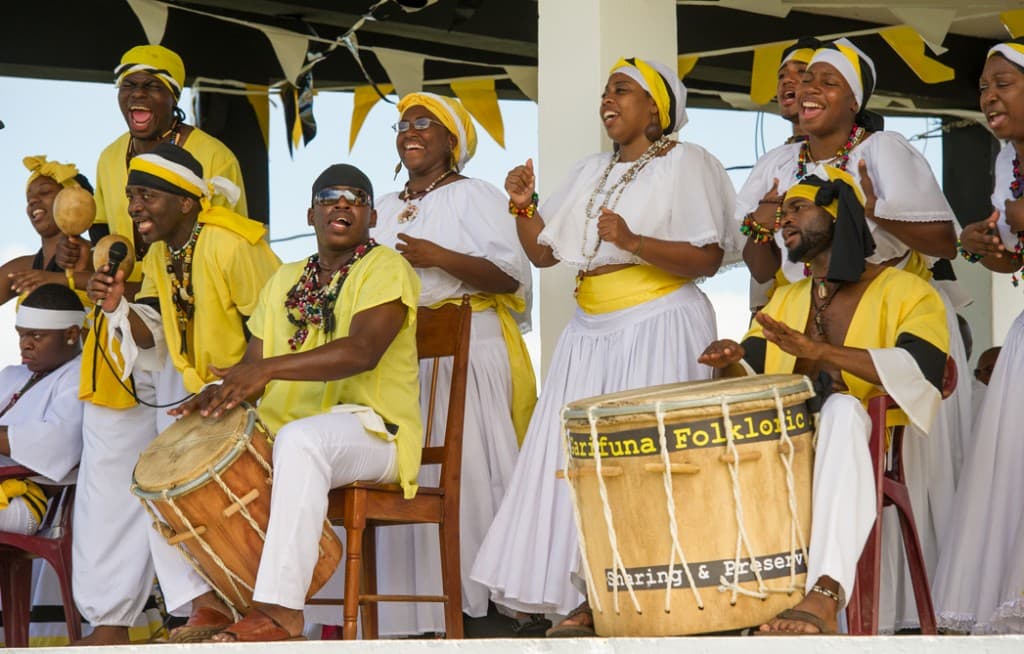 A lively group of performers in colorful costumes plays drums and sings together on stage.