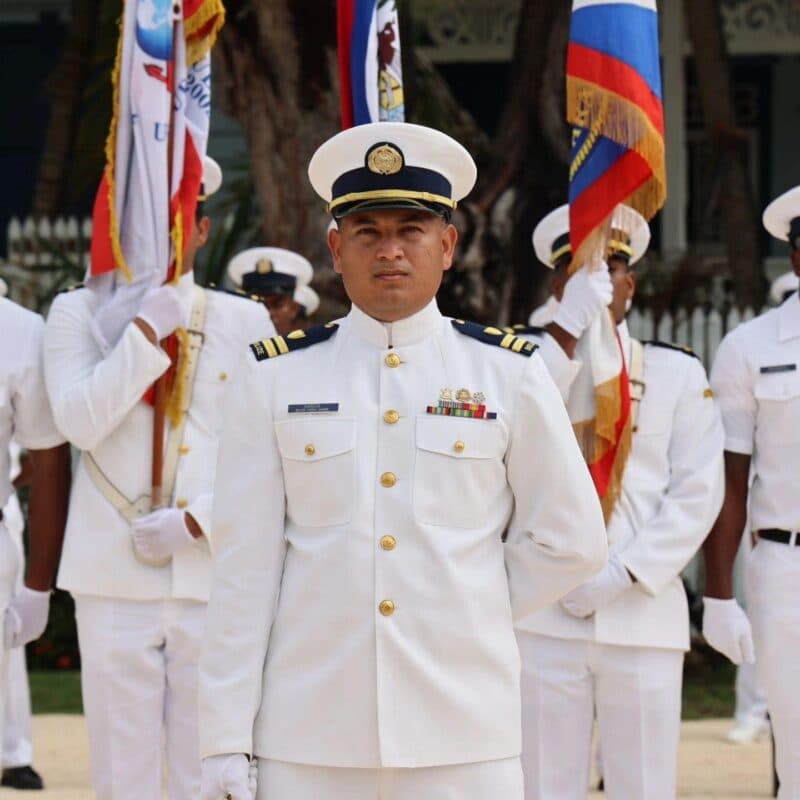 A naval officer stands at attention in white uniform, flanked by others holding flags.