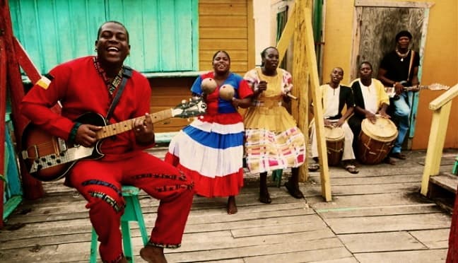 A group of musicians in colorful attire joyfully performing with guitars and drums on a wooden porch.
