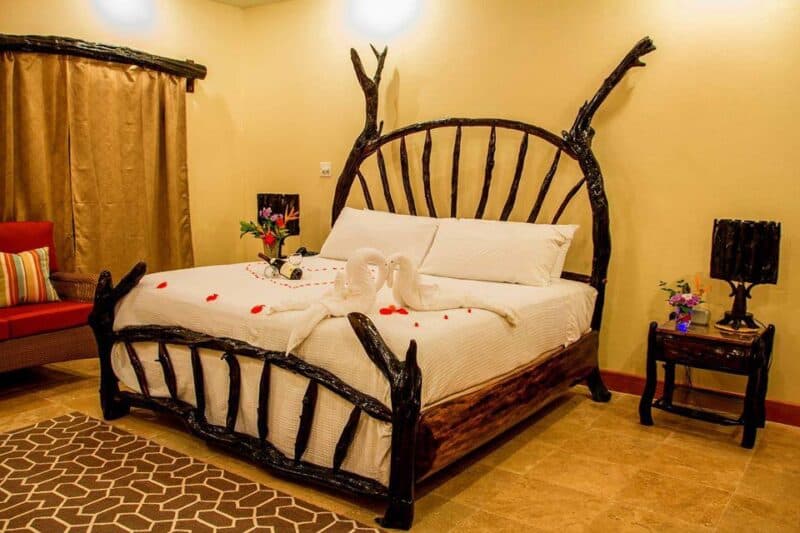 A rustic wooden bed with an artistic headboard, adorned with white linens and decorative rose petals, in a cozy room.