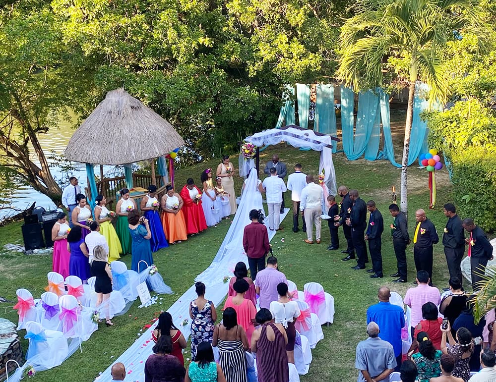 An outdoor wedding ceremony taking place near a river, with guests in colorful attire and a decorated arch.