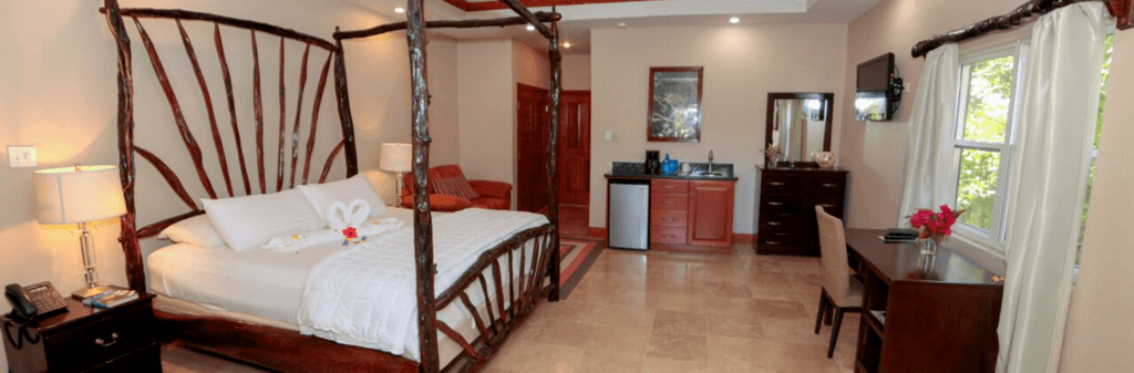 Spacious hotel room featuring a wooden four-poster bed, kitchenette, and seating area.
