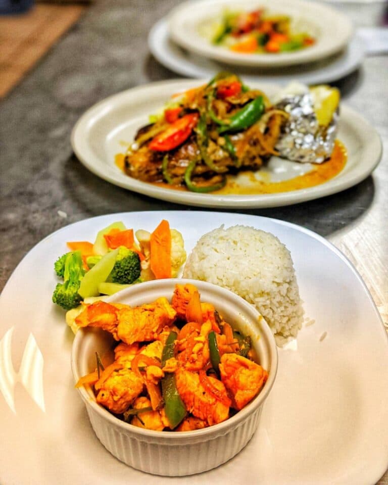 A plate of sautéed chicken with mixed vegetables and rice, with a second dish of grilled items in the background.