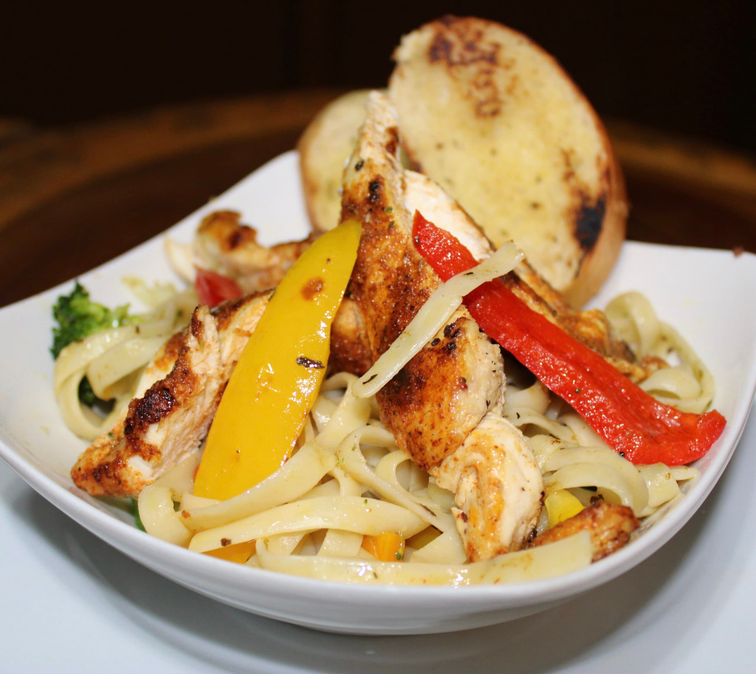 A bowl of fettuccine pasta topped with grilled chicken, colorful bell peppers, and accompanied by a piece of bread.