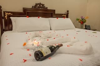 A romantic hotel room featuring a neatly made bed adorned with rose petals, a bottle of champagne, and elegantly arranged towels.