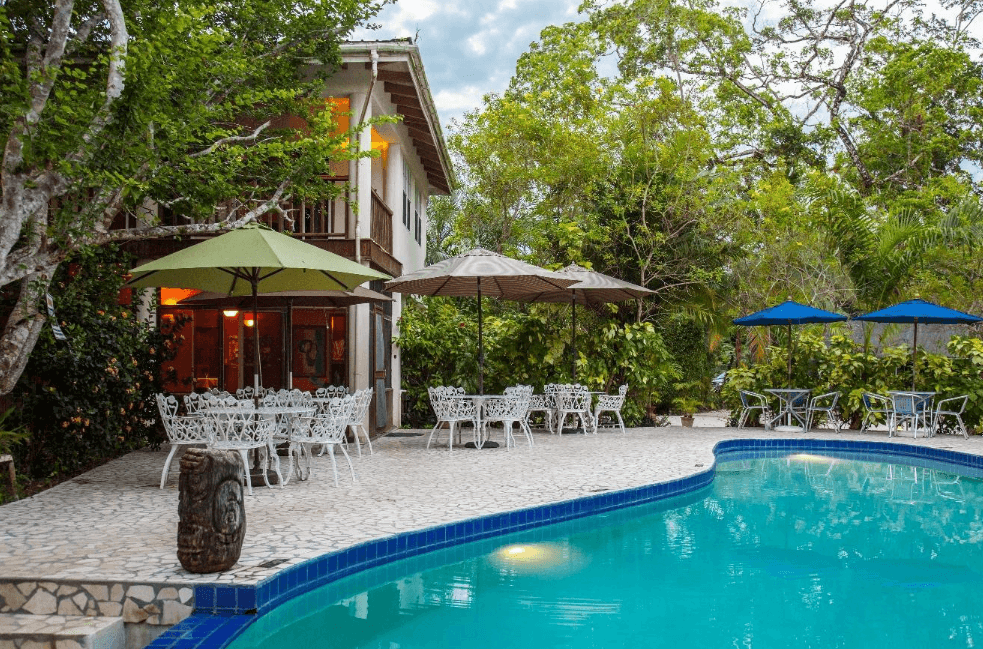 A tranquil outdoor area featuring a pool, shaded seating, and a nearby house surrounded by lush greenery.