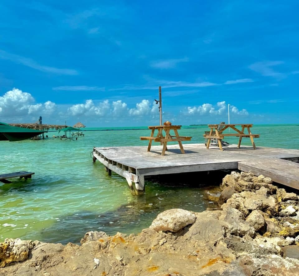A wooden dock with picnic tables extends over calm, turquoise water under a bright blue sky with scattered clouds.