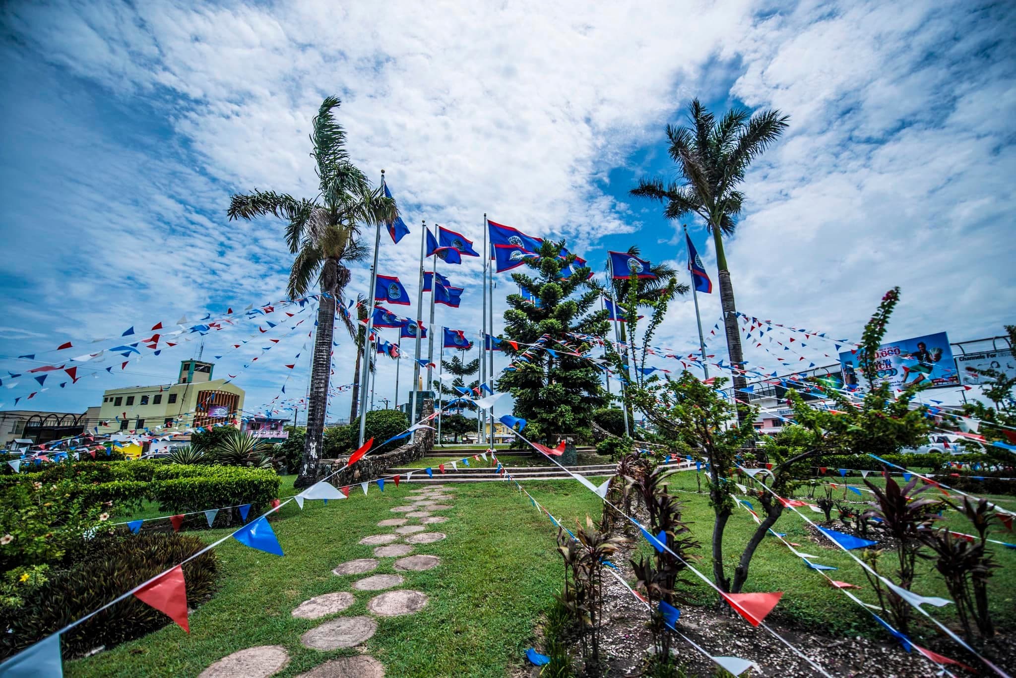 A vibrant outdoor area adorned with colorful flags and bunting, surrounded by palm trees and lush greenery under a cloudy sky.