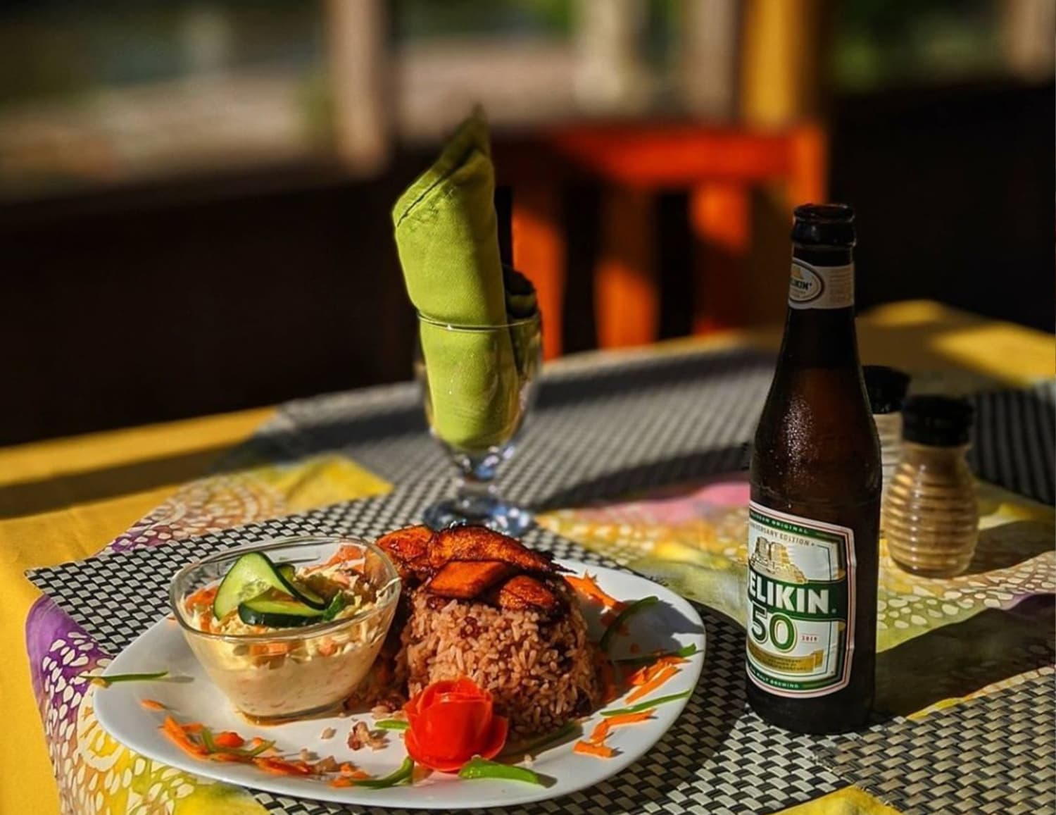 A plate of rice with plantains and coleslaw, accompanied by a bottle of beer and a green napkin on a colorful table.