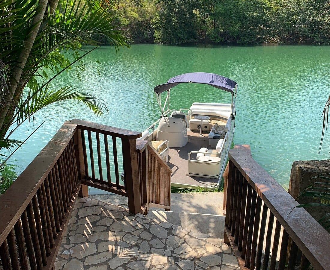 A wooden staircase leads down to a pontoon boat docked on a tranquil, green lake surrounded by lush vegetation.