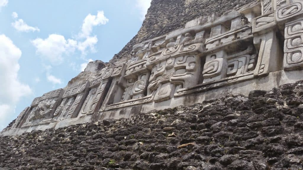Ancient Mayan carvings adorn a stone wall against a cloudy blue sky.