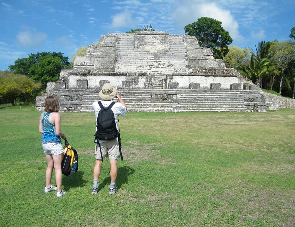 Two tourists admire a large ancient pyramid in a lush, green landscape.