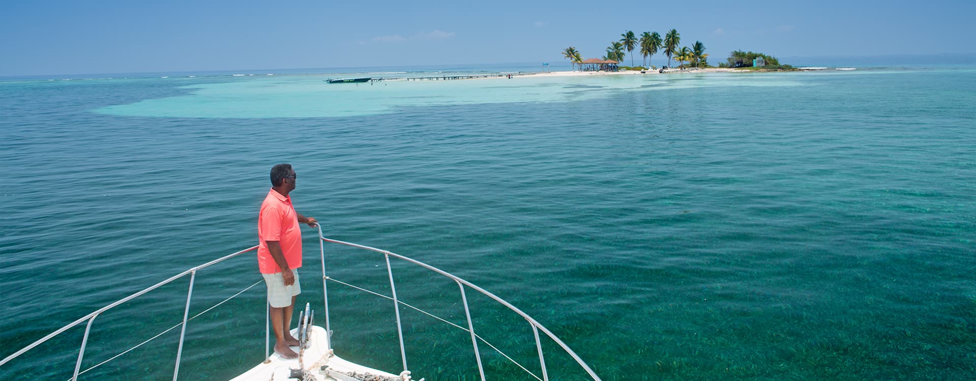A man stands at the bow of a boat, gazing out at a tropical island in clear blue waters.
