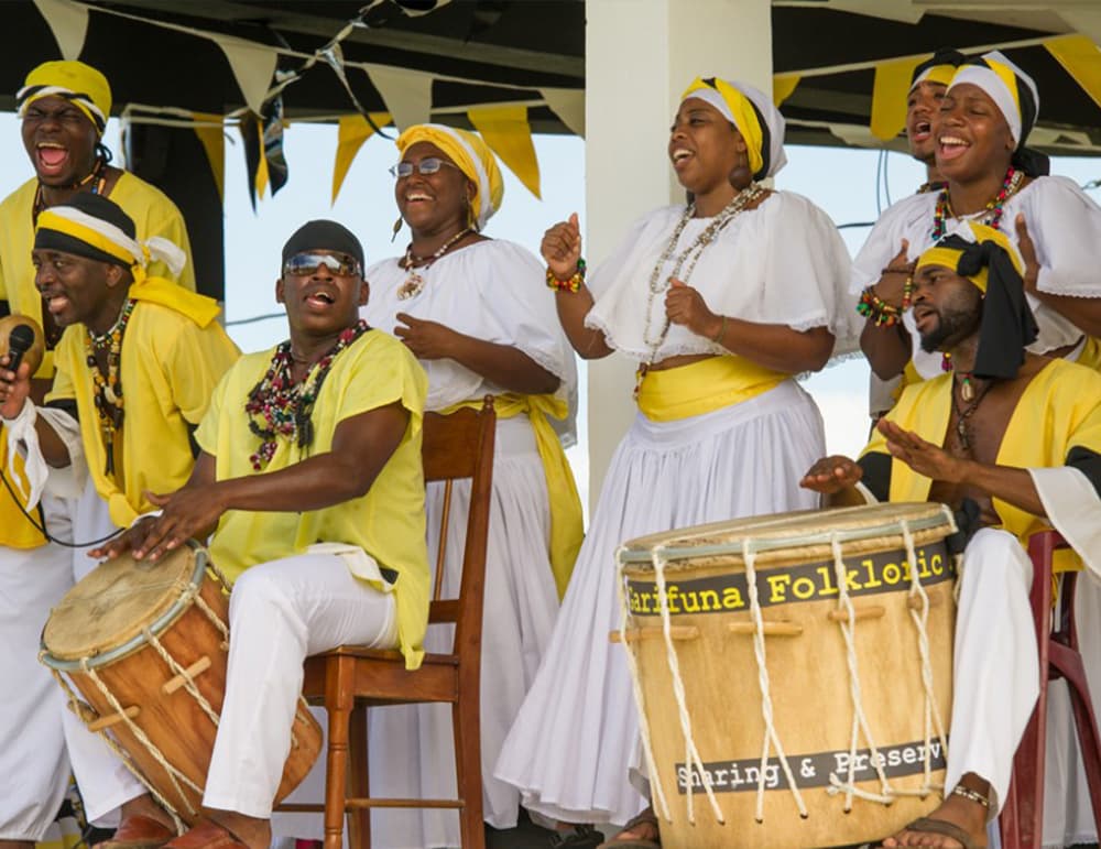 A group of performers in yellow and white clothing sings and plays traditional drums at a lively cultural event.