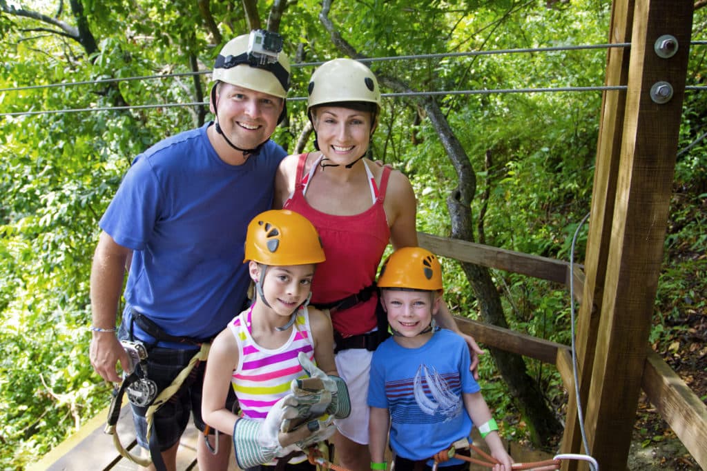 A family of four, wearing safety helmets and harnesses, poses together on a zip-lining platform surrounded by lush greenery.