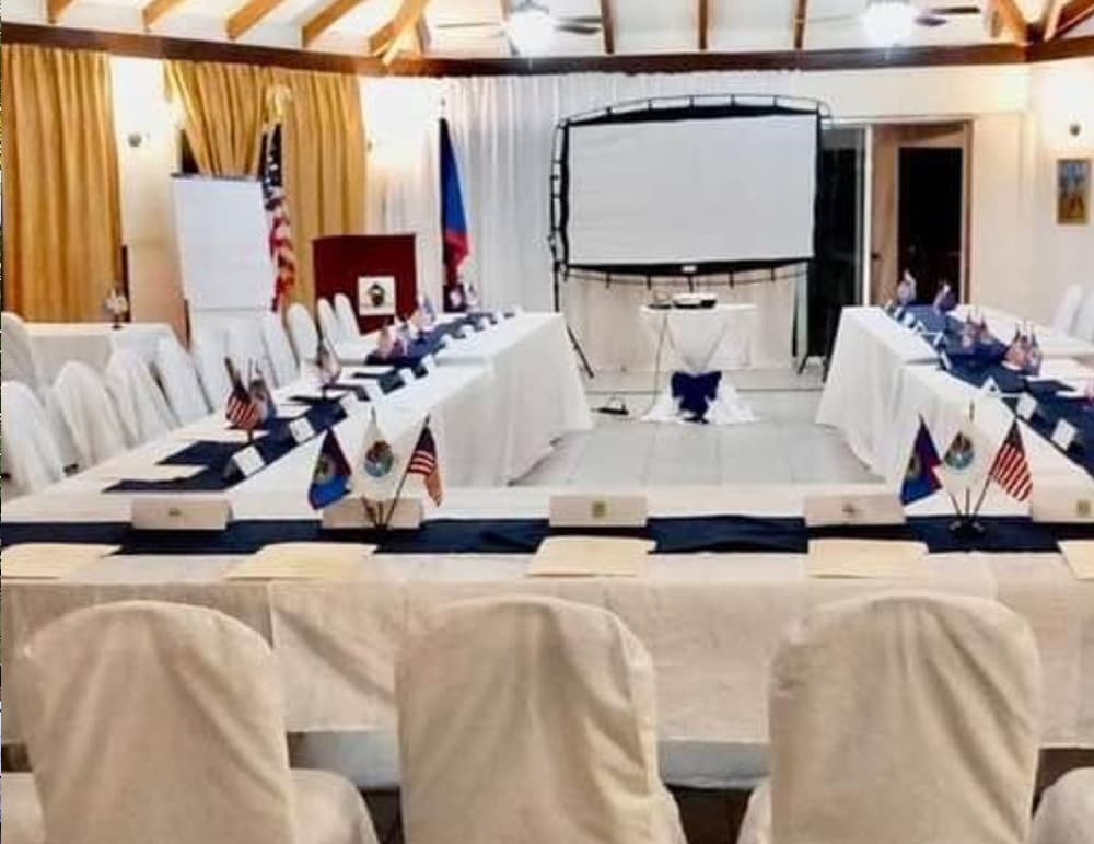 A conference room set up with tables, chairs, and flags of various countries, featuring a blank projector screen.