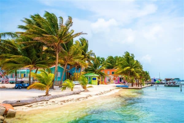 Colorful tropical buildings line a sandy beach with palm trees beside clear water.
