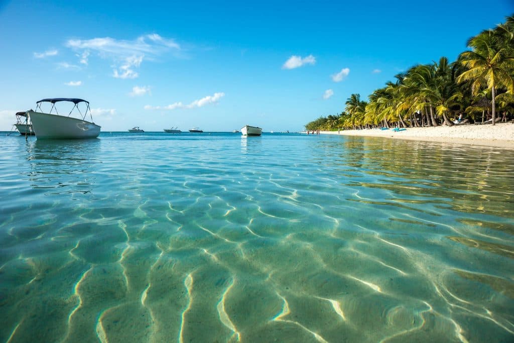 A serene beach scene with clear turquoise water, palm trees, and boats in the distance under a blue sky.