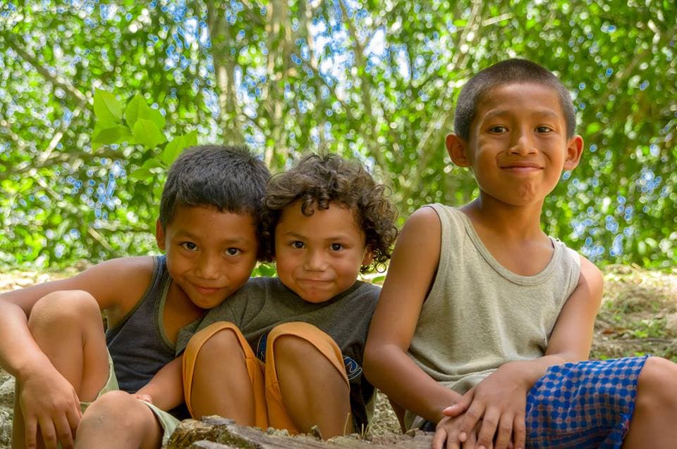 Three children sitting together outdoors, smiling amid lush greenery.