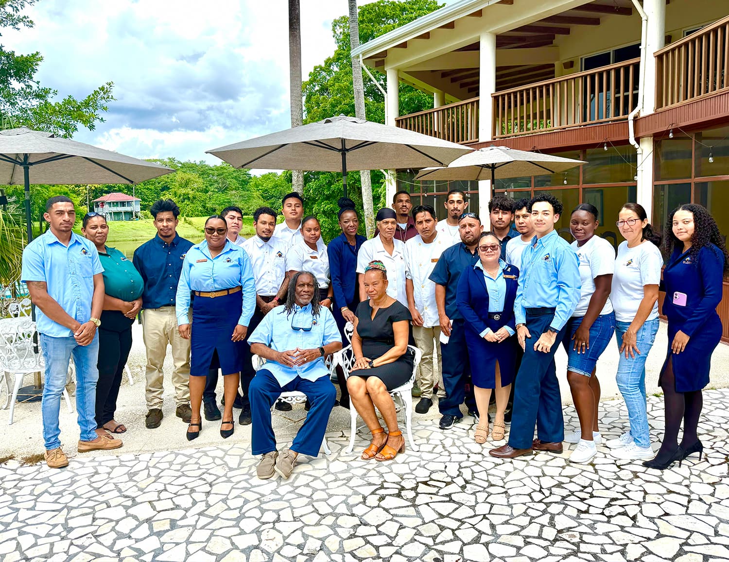 A diverse group of employees poses outdoors in front of a building, with umbrellas providing shade.