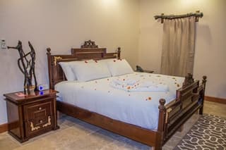 A romantic bedroom featuring a wooden bed with blue linens, rose petals scattered, and a bedside table.