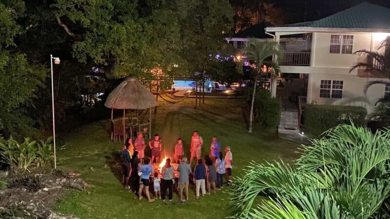 A group of people gathers around a campfire at night in a lush green setting with nearby buildings and a pool.