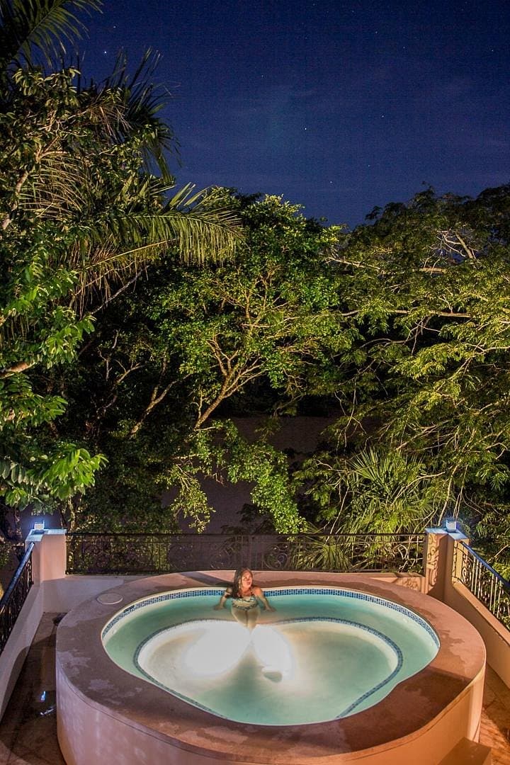 A person relaxes in a circular hot tub surrounded by lush greenery under a starry night sky.