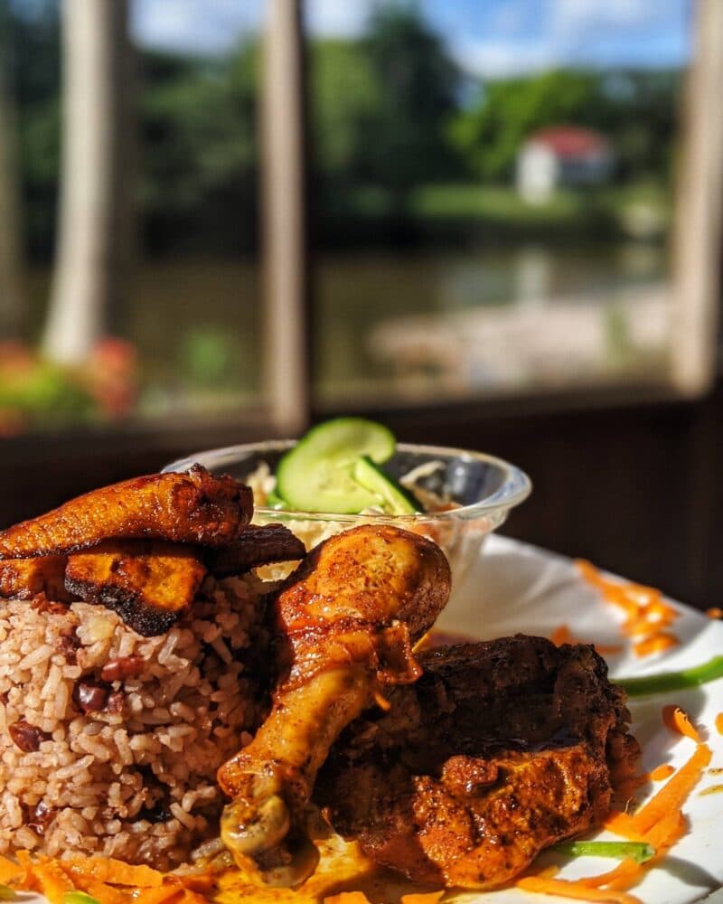 A plate of seasoned chicken, rice and beans, fried plantains, and a side of salad with cucumbers.