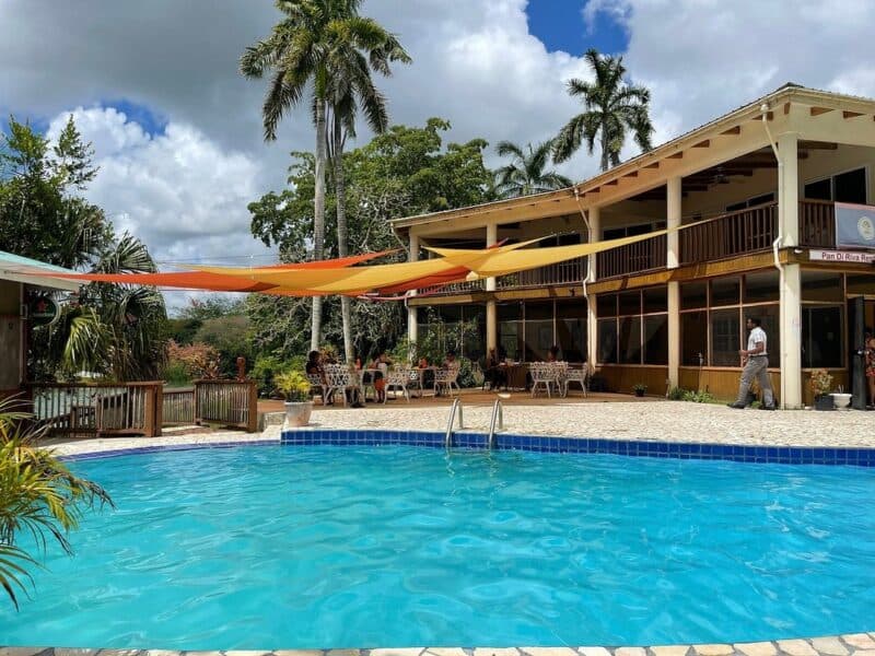 A clear blue pool surrounded by palm trees and a partially shaded outdoor restaurant.