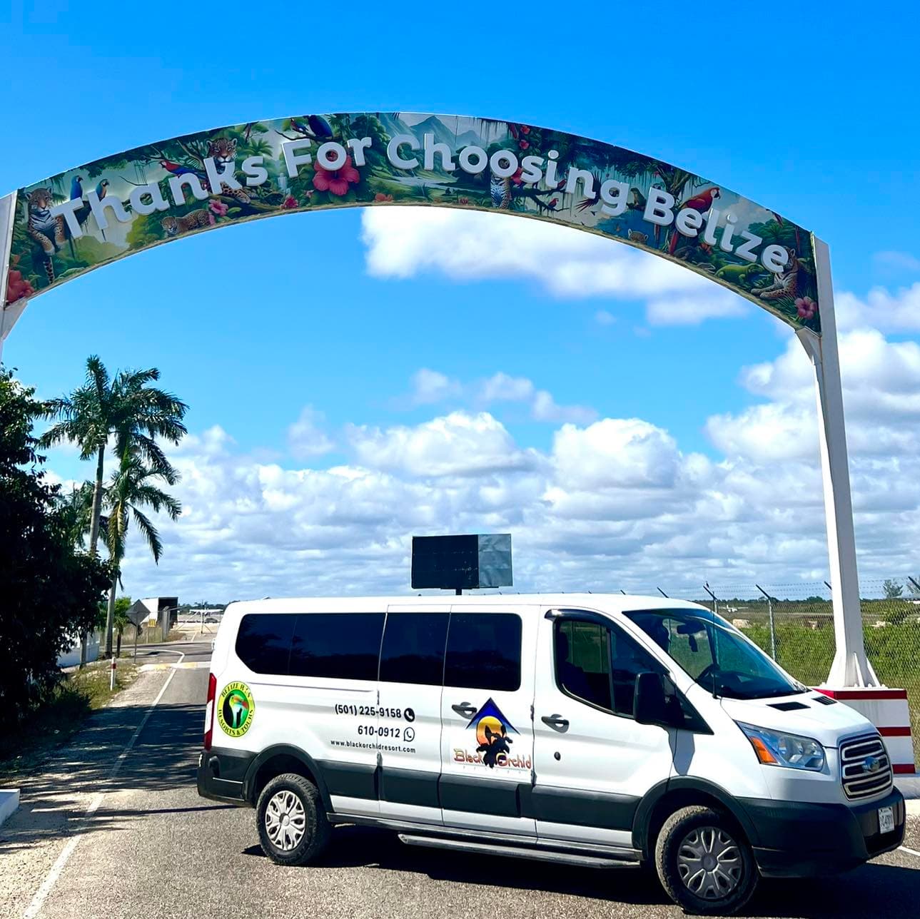 A white van parked under an archway that reads "Thanks For Choosing Belize" against a blue sky and palm trees.