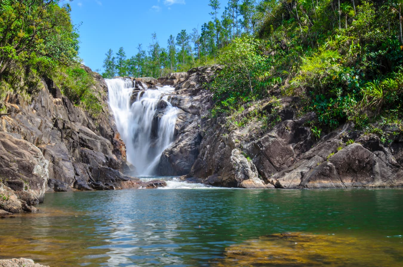 A tranquil waterfall cascades into a green pool, surrounded by lush foliage and rocky terrain.