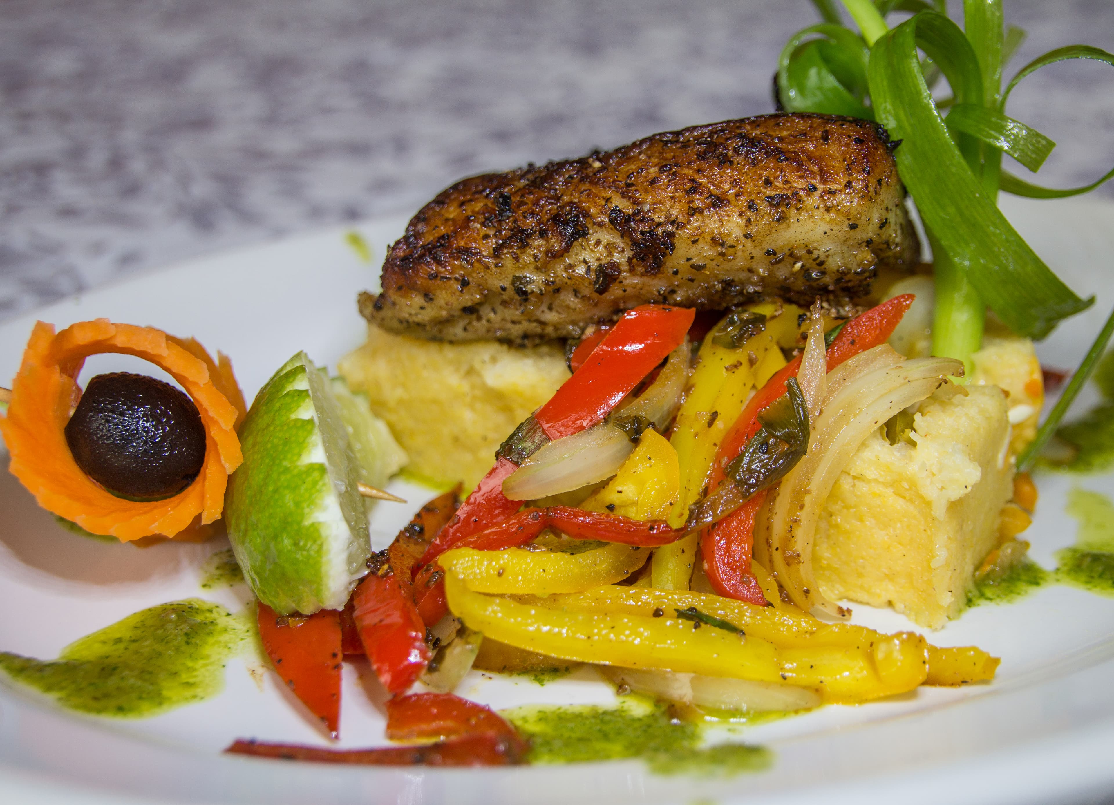 A plate of sautéed vegetables and seasoning topped with a seasoned fish fillet, garnished with lime and a carrot decoration.