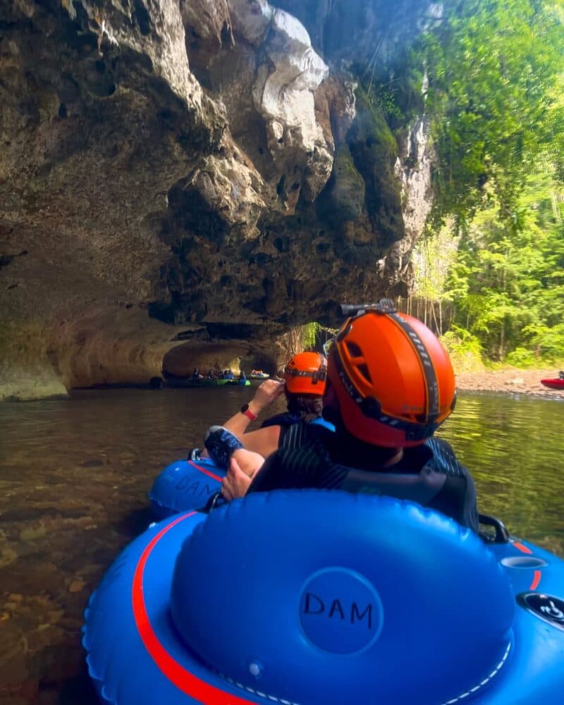 A group of people in inflatable rafts navigates through a cave entrance surrounded by lush greenery.