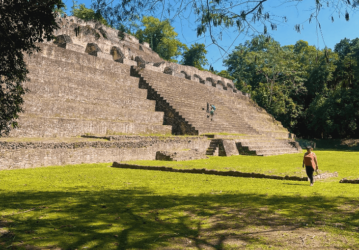 A person walks through a grassy area towards a large stone pyramid surrounded by trees.
