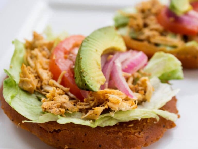Two tostadas topped with shredded chicken, lettuce, tomato, avocado, and sliced onion.