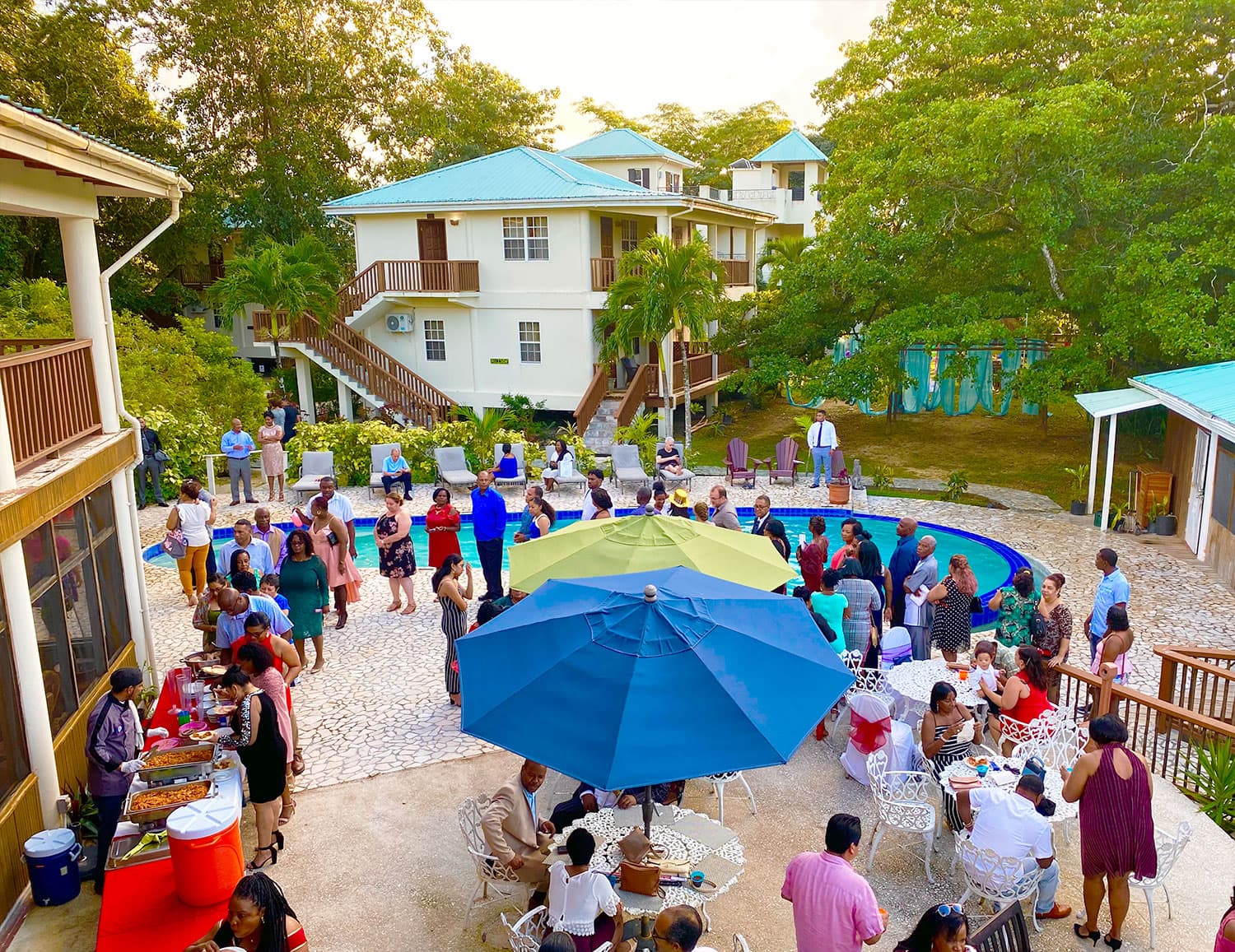 A lively outdoor gathering around a pool, with numerous guests mingling under colorful umbrellas and surrounded by trees and buildings.