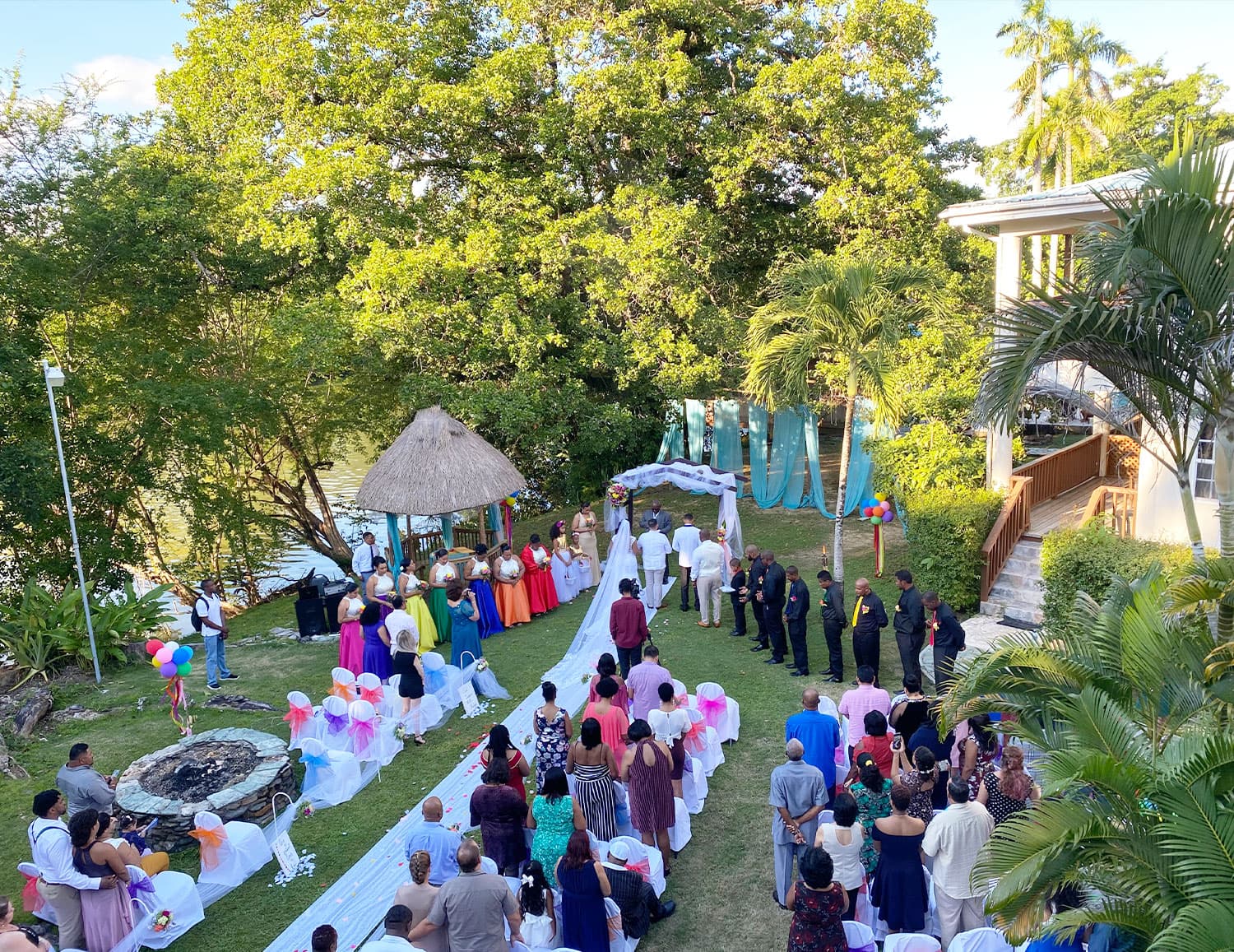 A wedding ceremony taking place outdoors with guests seated in colorful attire surrounded by greenery.