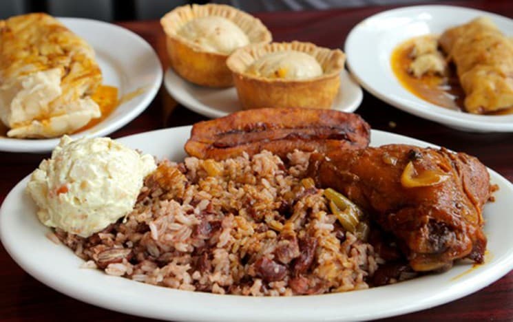 A plate of rice and beans, fried plantains, chicken with sauce, and a side of coleslaw, accompanied by two small servings of dessert.