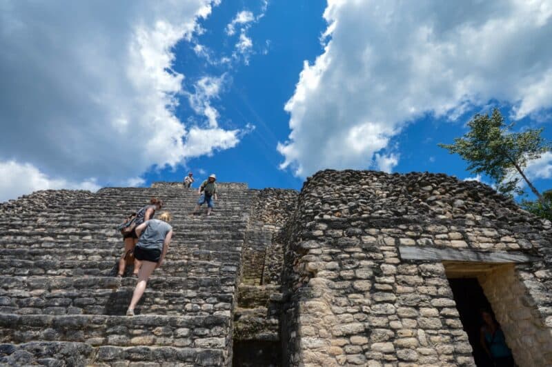 Visitors ascend stone steps of a Mayan ruin under a blue sky with scattered clouds.