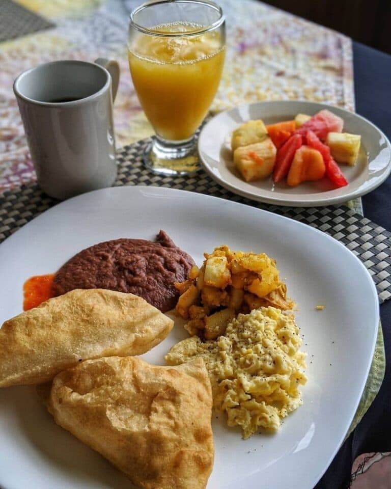 A plate of breakfast featuring two fried pastries, scrambled eggs, potatoes, refried beans with salsa, a side of mixed fruit, a cup of coffee, and orange juice.