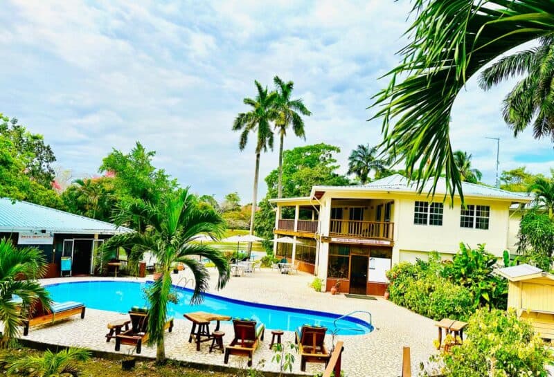 A tropical resort featuring a swimming pool surrounded by palm trees and lounge chairs, with a two-story building in the background.