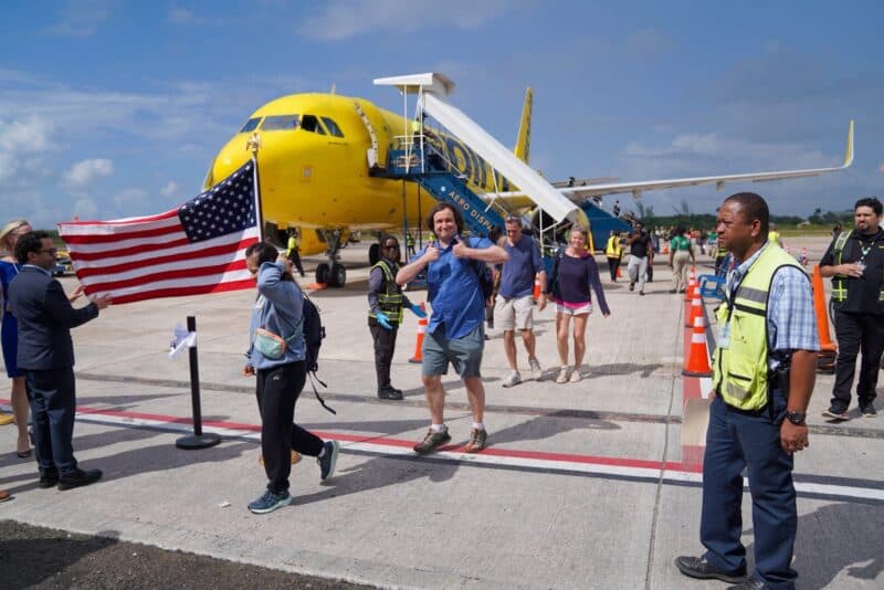Passengers disembark from a yellow airplane onto a sunny tarmac, with one man cheerfully waving a United States flag.