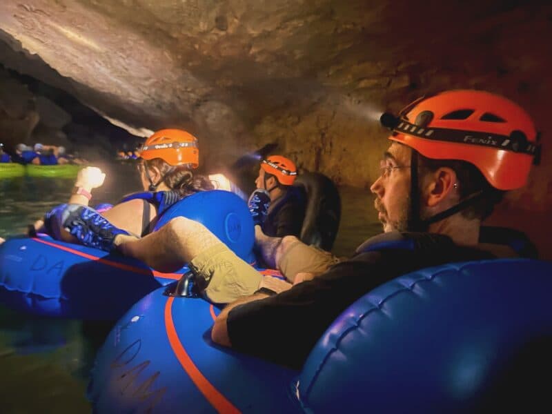 A group of people in helmets floats on blue inner tubes in a dark cave, illuminated by headlamps.