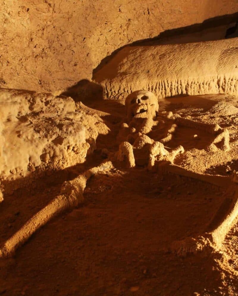 A skeleton partially buried in sand within a cave.