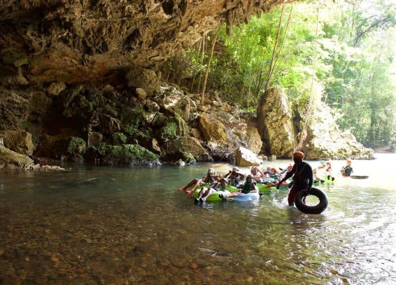 A group of people in kayaks paddles in a calm river surrounded by lush greenery and rocky cliffs.