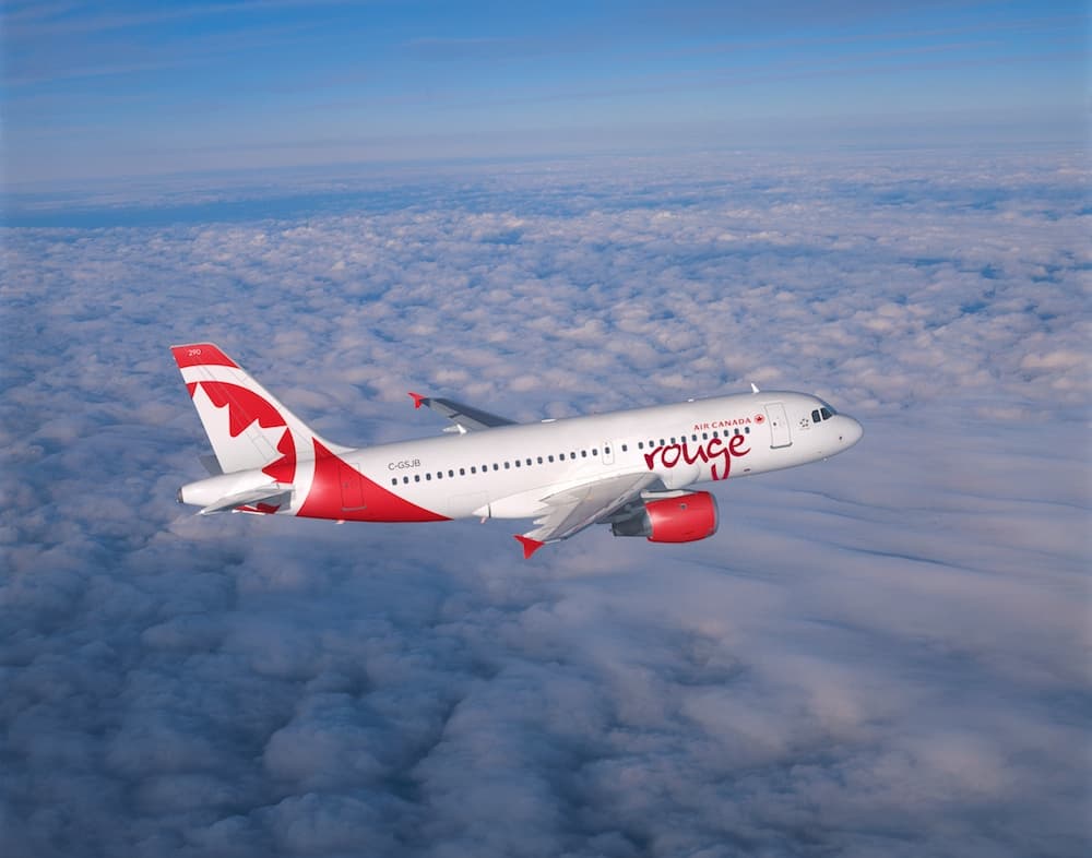 An Air Canada Rouge airplane flying above fluffy clouds against a blue sky.